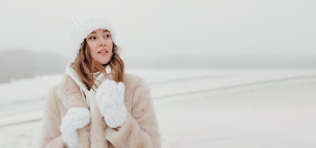 Foto donna caucasica con un cappello bianco di pelliccia e guanti che guardano di lato su uno sfondo di neve