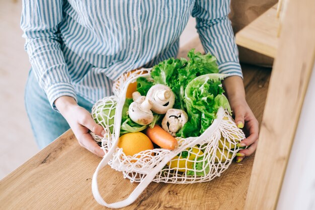 Caucasian woman hold eco net bag with fresh vegetables in modern kitchen.