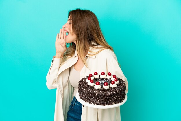 Foto donna caucasica che tiene la torta di compleanno isolata su fondo blu che grida con la bocca spalancata al lato