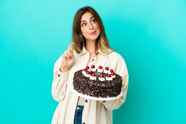 Caucasian woman holding birthday cake isolated on blue background with fingers crossing and wishing the best