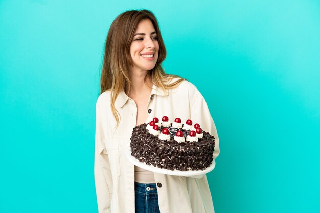 Caucasian woman holding birthday cake isolated on blue wall looking to the side and smiling