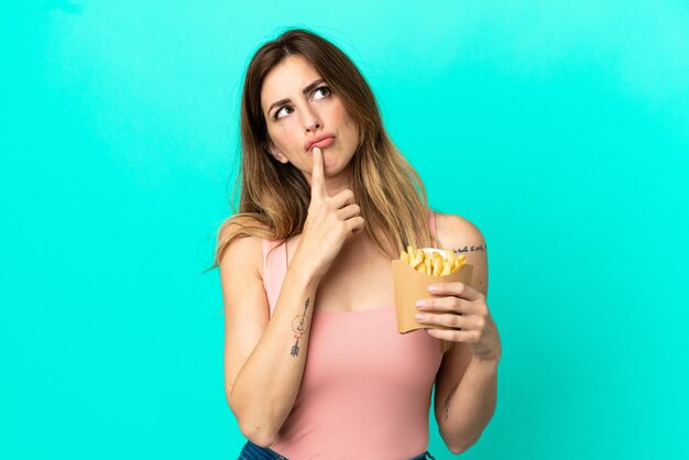 Caucasian woman holding fried chips isolated on blue background having doubts while looking up