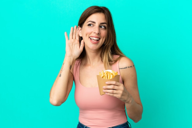 Caucasian woman holding fried chips isolated on blue background listening to something by putting hand on the ear