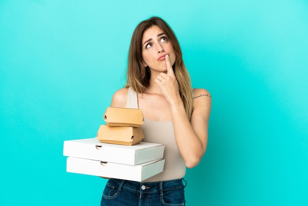 Caucasian woman holding pizzas and burger isolated on blue background having doubts while looking up