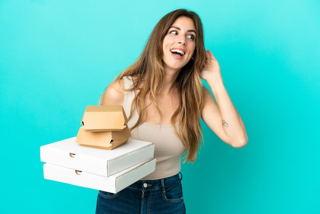 Caucasian woman holding pizzas and burger isolated on blue background listening to something by putting hand on the ear