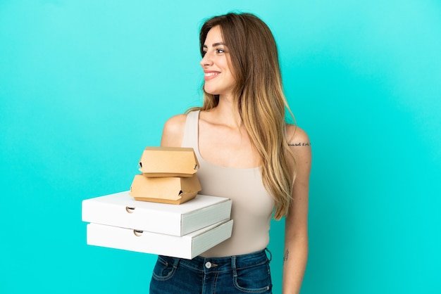 Caucasian woman holding pizzas and burger isolated on blue background looking side