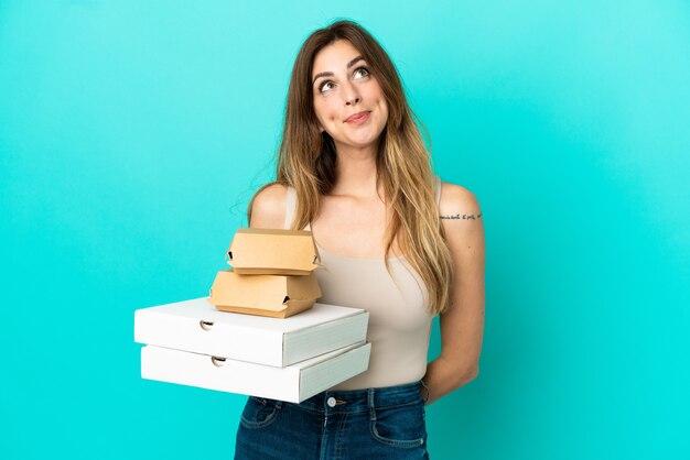 Caucasian woman holding pizzas and burger isolated on blue background and looking up