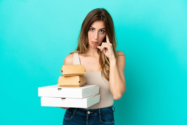 Caucasian woman holding pizzas and burger isolated on blue background thinking an idea