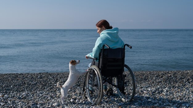 Caucasian woman in a hoodie in a wheelchair with a dog on the seashore