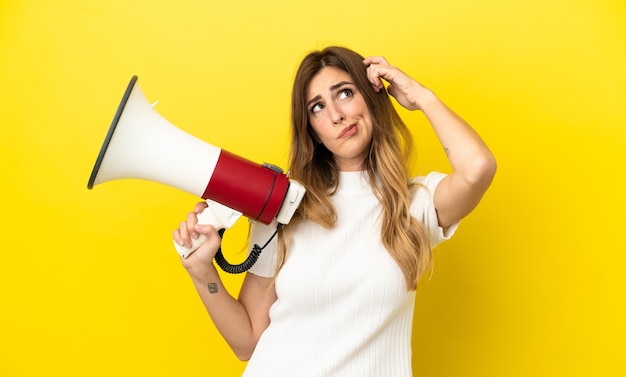 Caucasian woman isolated on yellow background holding a megaphone and having doubts