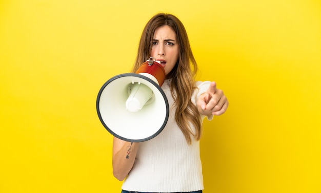 Caucasian woman isolated on yellow background shouting through a megaphone to announce something while pointing to the front