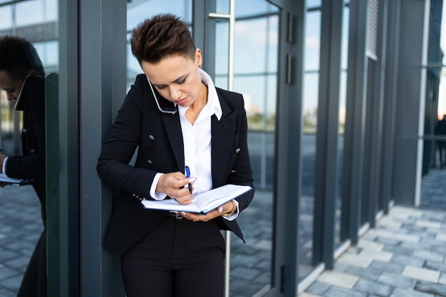 Photo caucasian woman in office clothes writes to notebook and waits colleague near the office building