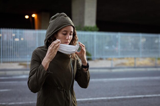 Caucasian woman out and about in the city streets during the day, wearing a hoodie, putting on a face mask against covid19 coronavirus