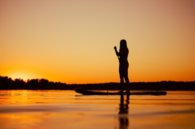 Caucasian woman pictured from low angle on paddle board with oar in hands looking at beautiful sunset covering water surface with orange color in evening Active lifestyle