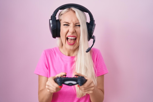 Caucasian woman playing video game holding controller angry and mad screaming frustrated and furious, shouting with anger. rage and aggressive concept.