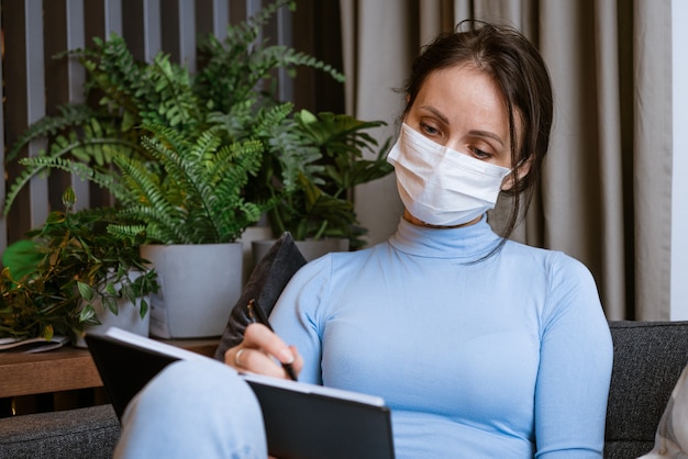 Photo caucasian woman in a protective mask sits on the couch with a notebook in her hand with a pensive look writes