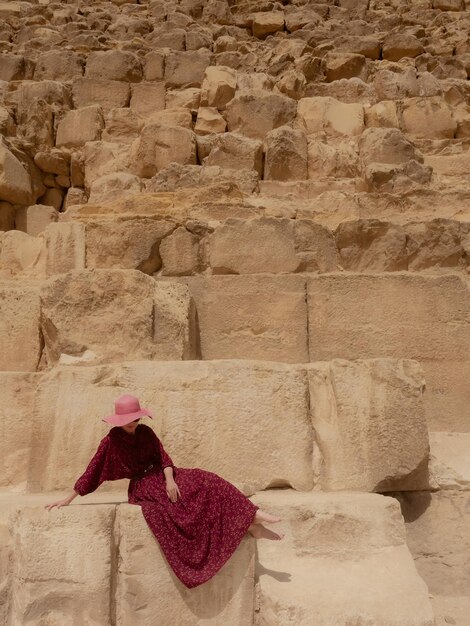 A caucasian woman in a red dress and hat sits beautifully khafre pyramid in giza