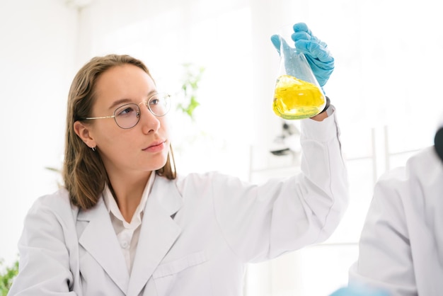 Caucasian woman scientist researcher wearing eye glasses\
shaking substance in the conical flask for analysis of liquids in\
the lab scientist working with a dropper and the conical flask