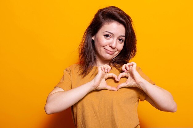 Caucasian woman showing heart shape with fingers at camera.
portrait of romantic person doing love sign and symbol with hands,
showing affection and emotion over orange background.