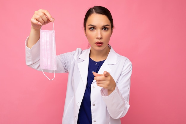 Caucasian woman showing protection face mask isolated on pink background and pointing finger at