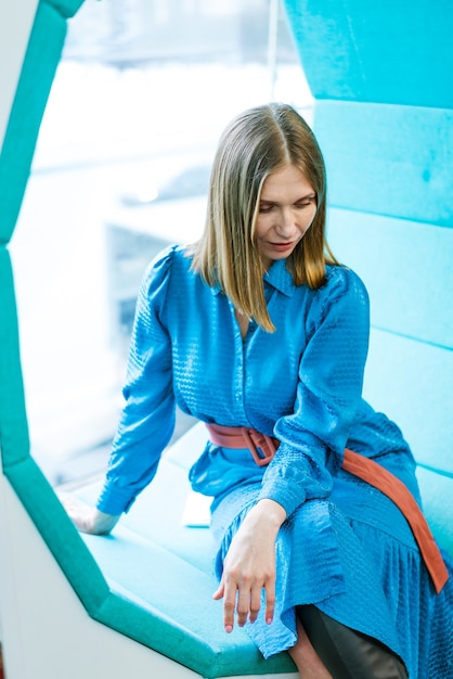 Caucasian woman sitting by window on bench in shape of circle in blue dress on the background of the...