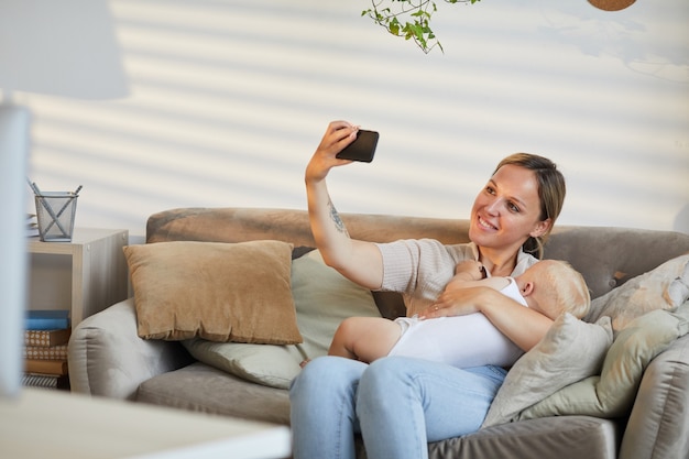 Caucasian woman sitting on sofa taking selfie photo on smartphone while cuddling her baby son in arms