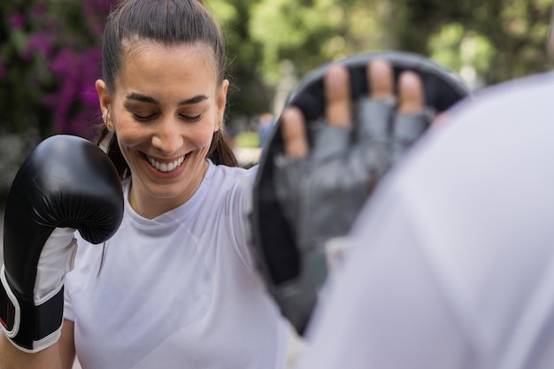 Кавказская женщина улыбается и занимается боксом на открытом воздухе. Она носит спортивную одежду и боксерские перчатки.