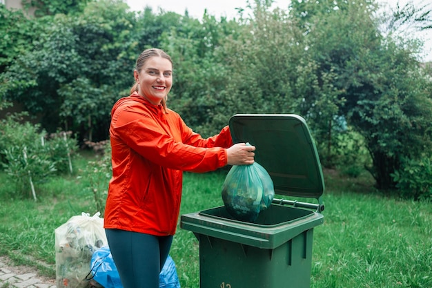 Foto donna caucasica che seleziona la spazzatura gettando un sacchetto di carta usato in un piccolo contenitore per il riciclaggio all'aperto vicino a un