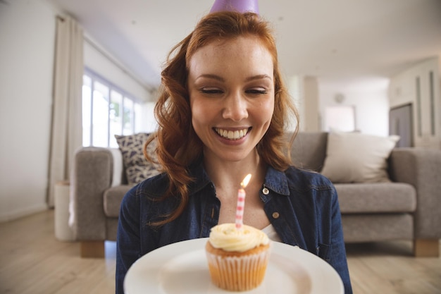 Caucasian woman spending time at home, in living room, smiling, celebrating and holding a cupcake with a candle. Social distancing during Covid 19 Coronavirus quarantine lockdown.