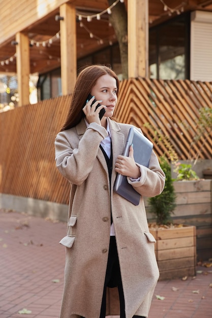 스마트폰 들고 노트북으로 전화하는 세련된 베이지색 코트에 백인 여자