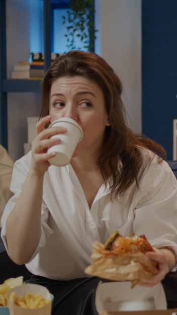 Caucasian woman taking bite from burger and sip of soda sitting\
on couch at tv. person eating takeaway food and drinking juice\
watching television. adult having unhealthy dinner at home