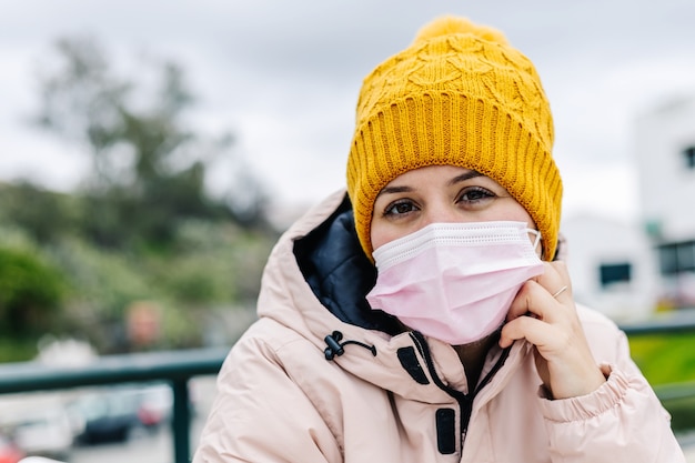 Photo caucasian woman up close looking at camera, wearing yellow wool cap, pensive with pink face mask