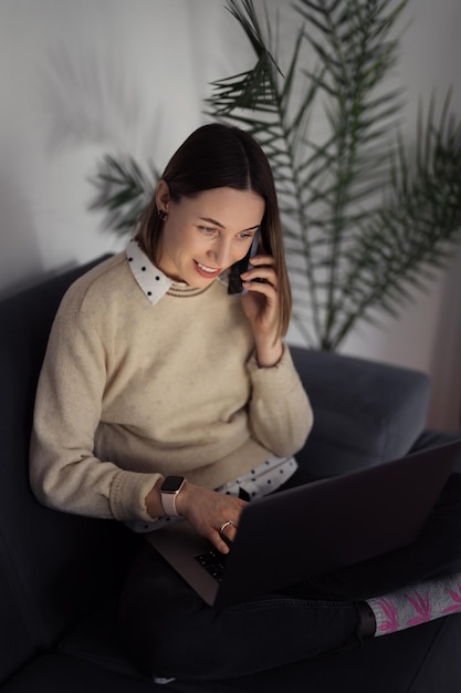 Photo caucasian woman using laptop while sitting on the sofa at home in the evening living room speaking on the phone