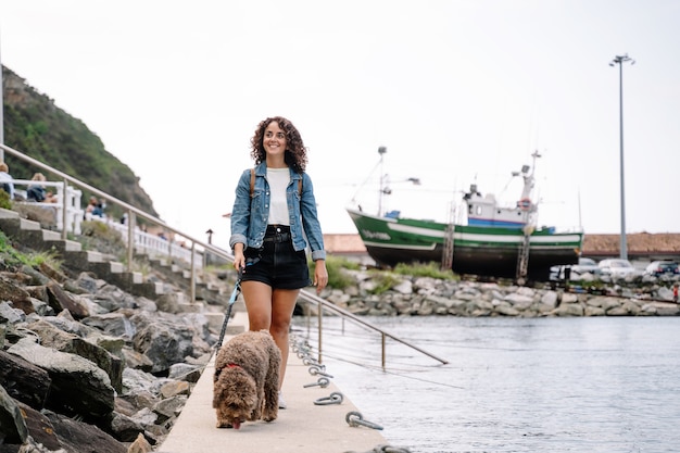 港の散歩を横切って彼女の水犬を歩いている白人女性。動物と一緒に旅行する女性の水平方向のビュー。動物と旅行のコンセプト。