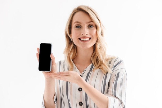 caucasian woman wearing casual clothes smiling at camera and holding smartphone isolated over white wall