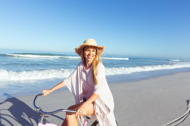 A Caucasian woman wearing a hat enjoying time at the beach on a sunny day, riding a bike, with sea in the background