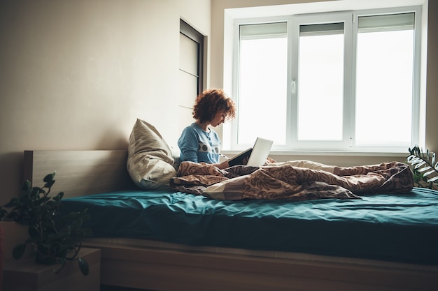 Donna caucasica con capelli ricci che legge un libro nel suo letto seduto vicino alla finestra con un computer