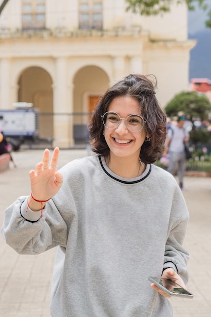 Caucasian woman with glasses waving and holding a smart phone in her hands.