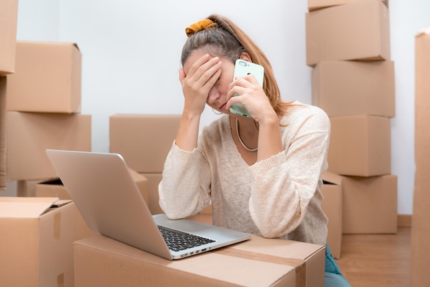 Caucasian woman with headache from moving to new home with laptop and talks to consultant on phone