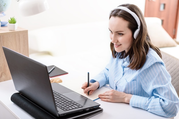 Caucasian woman with headphones writes in notebook, looks at laptop screen