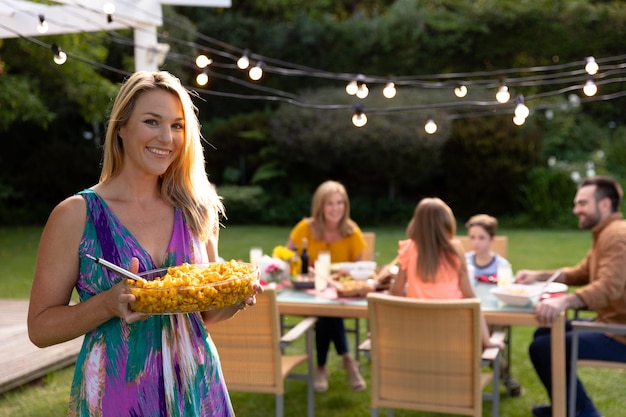 A Caucasian woman with long blonde hair standing in her garden holding a dish of food and smiling to camera, in the background her multi-generation family sitting down at a table for a meal outside