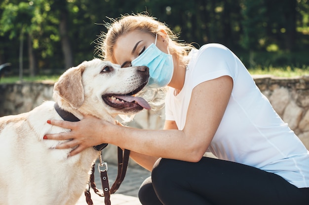 Caucasian woman with medical mask and blonde hair is embracing her labrador while walking in a park