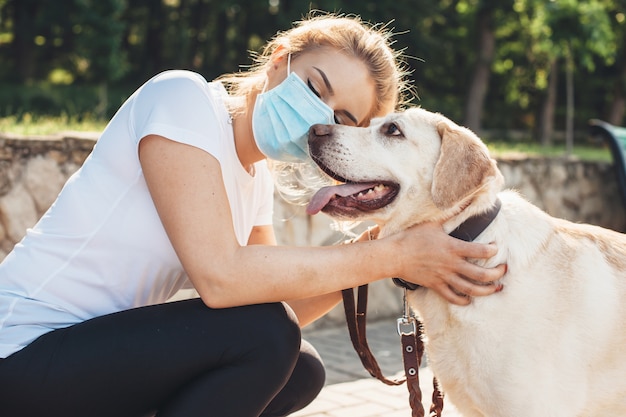 Photo caucasian woman with medical mask on face is embracing her golden retriever during a walk in park