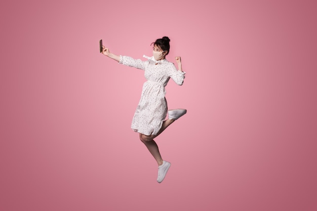 Кавказская женщина с медицинской маской носит платье и делает селфи по телефону, прыгая на розовой стене студии
