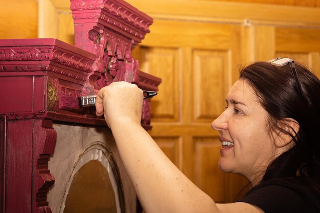 Кавказская женщина с улыбкой на лице красит деревянный орнаментированный шкаф в красном цвете большой кистью с дверями на заднем плане Повторное использование старых антикварных вещей Мастерская по ремонту домашней мебели