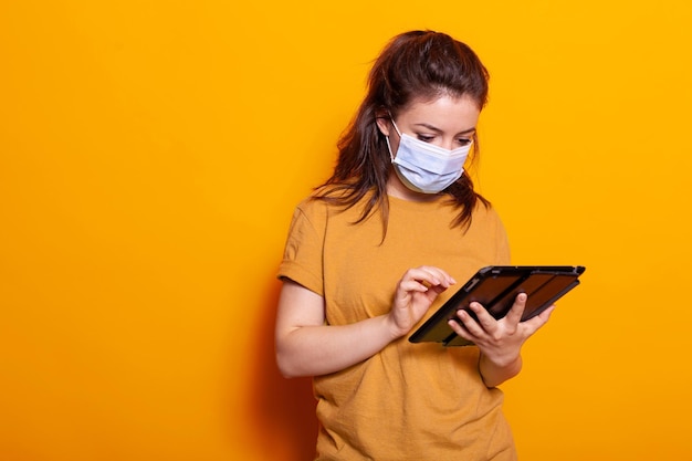 Caucasian woman working on display of digital tablet in studio. young adult wearing face mask and looking at modern gadget during coronavirus pandemic. adult over isolated background