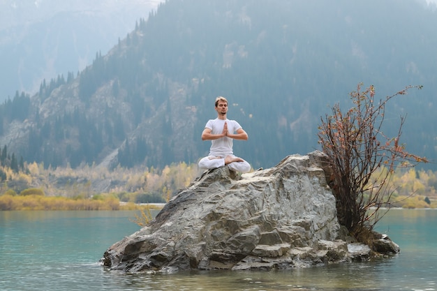 Caucasian yoga man in outdoor meditation sitting on lonely rock island of mountain lake.