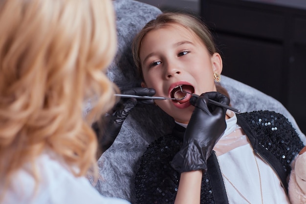 Caucasian young child girl sitting in medical chair while dentist fixing teeth at dental clinic using dental tools instruments, close-up photo of face. professional medical specialist.