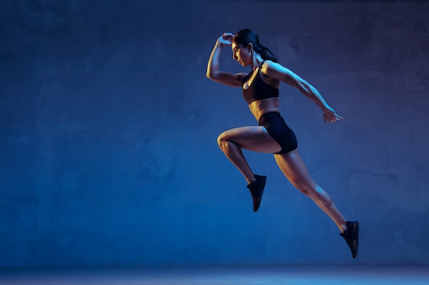 Foto caucasica giovane atleta femminile che pratica su sfondo blu studio in luce al neon. primo piano del modello sportivo che salta in alto, correndo. body building, stile di vita sano, concetto di bellezza e azione.