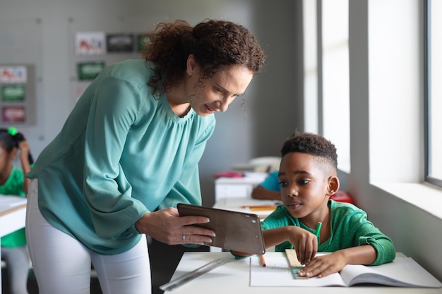 Photo caucasian young female teacher showing digital tablet to african american elementary boy in class